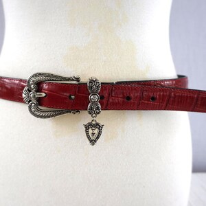 K&ouml;nnte beinhalten: Ein roter Lederg&uuml;rtel mit silberner Schnalle und Zierakzenten. Der G&uuml;rtel hat ein strukturiertes, krokodilartiges Muster. Die Schnalle ist verziert, mit einem herzf&ouml;rmigen Ende. Ein silberner Anh&auml;nger h&auml;ngt am G&uuml;rtel.