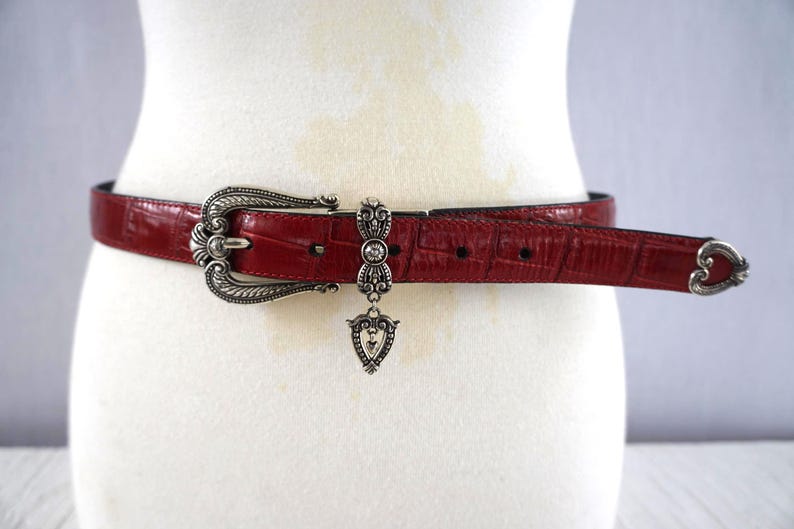 K&ouml;nnte beinhalten: Ein roter Lederg&uuml;rtel mit silberner Schnalle und Zierakzenten. Der G&uuml;rtel hat ein strukturiertes, krokodilartiges Muster. Die Schnalle ist verziert, mit einem herzf&ouml;rmigen Ende. Ein silberner Anh&auml;nger h&auml;ngt am G&uuml;rtel.