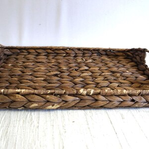 May include: Rectangular woven tray with built-in handles. The tray is made of a natural brown material, likely woven water hyacinth, and has a textured surface. The handles are curved and integrated into the tray's design. Ideal for serving or decorative use.
