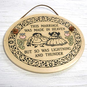 May include: Oval ceramic plaque with a brown leather strap for hanging. The plaque features a cartoon couple and the text "THIS MARRIAGE WAS MADE IN HEAVEN BUT SO WAS LIGHTNING AND THUNDER."