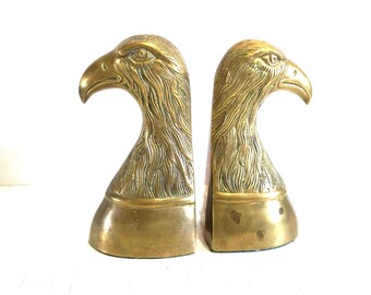 May include: A pair of polished brass bookends, each shaped as the head of an eagle. The metal features detailed engravings of feathers and sharp beaks, with the eagle heads facing in opposite directions, set upon smooth, curved bases.
