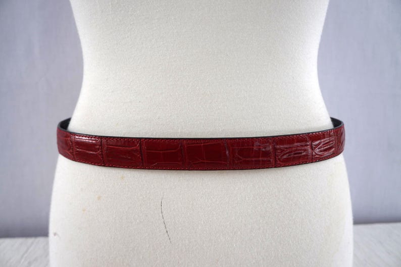 K&ouml;nnte beinhalten: Ein weinroter G&uuml;rtel mit Krokodilleder-Textur ist um eine Schaufensterpuppe gelegt. Der G&uuml;rtel ist schmal, mit einer dunklen Unterseite. Ein klassisches Accessoire.