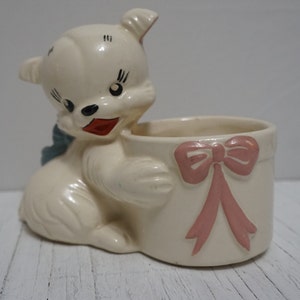 May include: White ceramic planter shaped like a dog holding a gift box with a pink bow. The dog has black eyes and a red mouth.