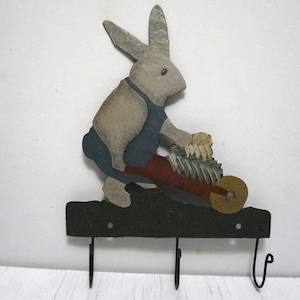 May include: A wooden wall-mounted key holder featuring a rabbit pushing a wheelbarrow. The rabbit is painted in shades of gray, beige, and blue, with a red wheelbarrow and green base. Three black hooks are attached below.