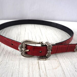K&ouml;nnte beinhalten: Roter Lederg&uuml;rtel mit silberfarbener Schnalle. Der G&uuml;rtel hat eine strukturierte Oberfl&auml;che und eine schwarze Unterseite. Die Schnalle hat ein verziertes Design. Ein stilvolles Accessoire.