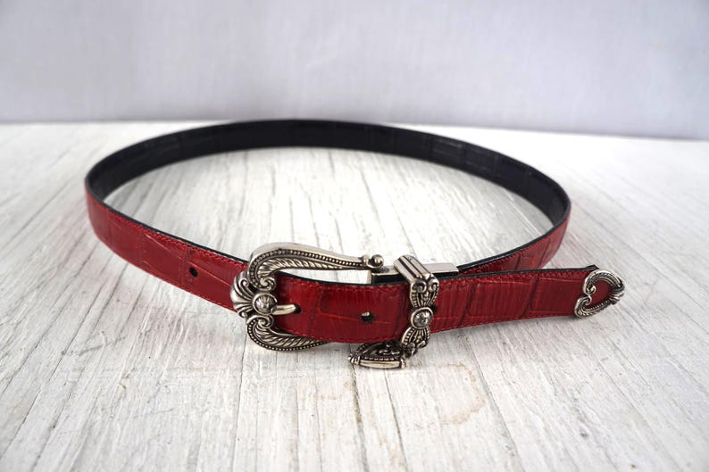 K&ouml;nnte beinhalten: Roter Lederg&uuml;rtel mit silberfarbener Schnalle. Der G&uuml;rtel hat eine strukturierte Oberfl&auml;che und eine schwarze Unterseite. Die Schnalle hat ein verziertes Design. Ein stilvolles Accessoire.