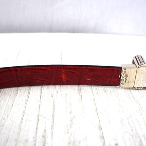 K&ouml;nnte beinhalten: Ein roter Lederg&uuml;rtel mit Krokodiltextur. Der G&uuml;rtel hat eine silberfarbene Schnalle mit dekorativen Details. Die Schnalle ist rechteckig und hat ein Scharnierdesign. Der G&uuml;rtel liegt auf einer wei&szlig;en Holzoberfl&auml;che.