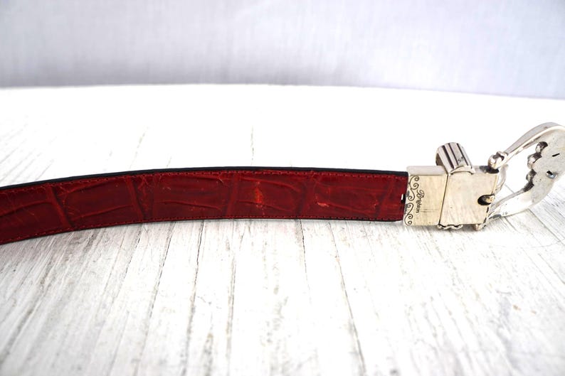 K&ouml;nnte beinhalten: Ein roter Lederg&uuml;rtel mit Krokodiltextur. Der G&uuml;rtel hat eine silberfarbene Schnalle mit dekorativen Details. Die Schnalle ist rechteckig und hat ein Scharnierdesign. Der G&uuml;rtel liegt auf einer wei&szlig;en Holzoberfl&auml;che.