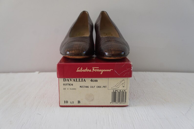 May include: A pair of brown leather women's shoes with a crocodile print. The shoes are in a box with the brand name Salvatore Ferragamo and the model name Davallia. The box also includes the size 10 1/2 B and the heel height, 4 cm.