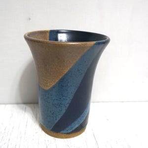 以下が含まれることがあります： 広がった縁と先細りの本体を持つ陶器製のタンブラー。外側の表面は、マットなベージュの粘土、斑点のあるライトブルーの釉薬、そして光沢のあるダークネイビーブルーの釉薬のセクションで構成された、対角線のカラーブロックパターンが特徴です。