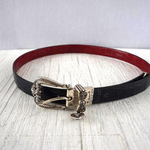 K&ouml;nnte beinhalten: Ein schwarzer G&uuml;rtel mit roter Innenseite und silberner Schnalle. Der G&uuml;rtel hat ein strukturiertes, krokodilartiges Muster. Die Schnalle ist mit einem dekorativen Design verziert.