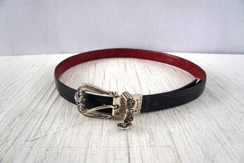 K&ouml;nnte beinhalten: Ein schwarzer G&uuml;rtel mit roter Innenseite und silberner Schnalle. Der G&uuml;rtel hat ein strukturiertes, krokodilartiges Muster. Die Schnalle ist mit einem dekorativen Design verziert.