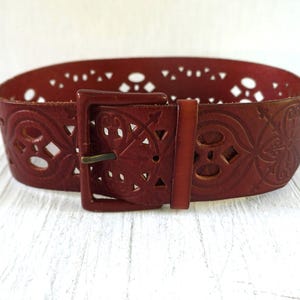 May include: A wide, reddish-brown leather belt with intricate cut-out designs and a rectangular buckle. The belt features a decorative pattern and a brass-colored buckle. The belt is approximately 3 inches wide.