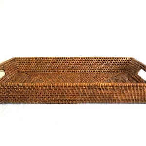 May include: Rectangular woven rattan tray in a warm brown hue. The tray features built-in handles on each end for easy carrying. The woven design creates a textured surface, suitable for serving or decorative purposes.