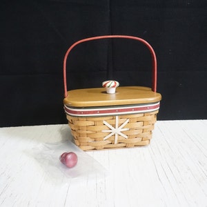 May include: A small, brown wicker basket with a red handle and a white snowflake design. The basket has a wooden lid with a red and green candy cane knob.