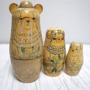 May include: A set of three wooden nesting dolls, each painted with a bear design. The largest doll has a bear head and ears. The dolls are painted in shades of beige, with floral and decorative accents in green and yellow.