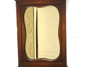 May include: A small, antique wooden wall mirror with a drawer. The mirror has a unique, curved shape and is framed by a dark brown wooden border. The mirror has a small shelf and a drawer with two round knobs.
