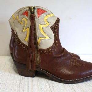 K&ouml;nnte beinhalten: Braune Leder-Cowgirl-Stiefel mit einem wei&szlig;en und goldenen Muster. Die Stiefel haben einen Rei&szlig;verschluss an der Seite und ein Fransen-Detail.