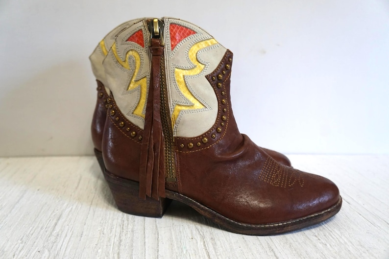 K&ouml;nnte beinhalten: Braune Leder-Cowgirl-Stiefel mit einem wei&szlig;en und goldenen Muster. Die Stiefel haben einen Rei&szlig;verschluss an der Seite und ein Fransen-Detail.