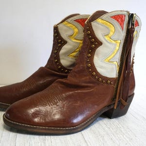 K&ouml;nnte beinhalten: Braune Lederstiefel mit einem wei&szlig;en und gelben Muster. Die Stiefel haben eine spitze Zehenpartie und einen niedrigen Absatz. Die Stiefel sind mit goldenen Nieten verziert.