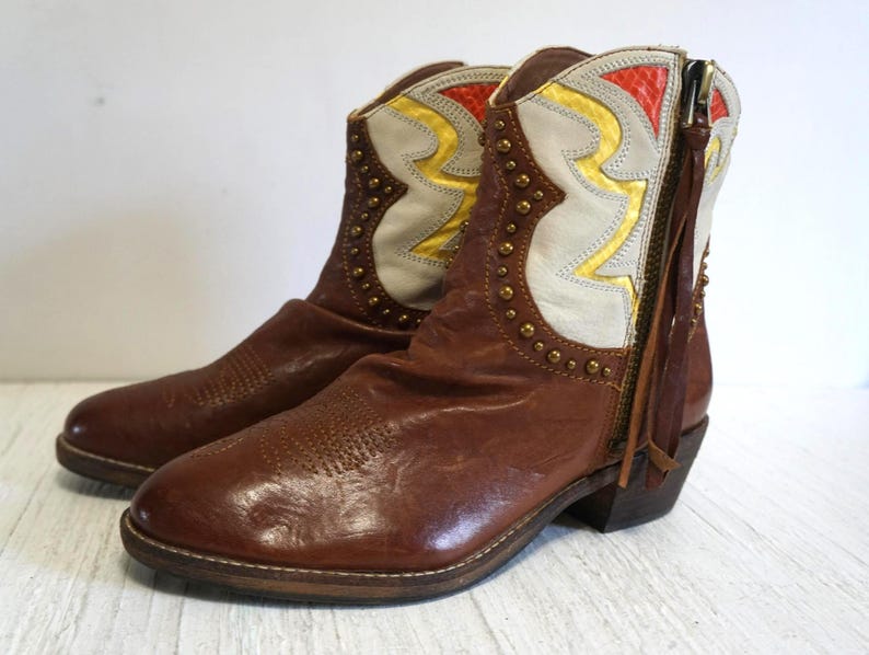 K&ouml;nnte beinhalten: Braune Lederstiefel mit einem wei&szlig;en und gelben Muster. Die Stiefel haben eine spitze Zehenpartie und einen niedrigen Absatz. Die Stiefel sind mit goldenen Nieten verziert.