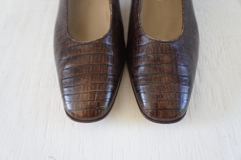 May include: A pair of brown leather flats with a crocodile-embossed pattern. The shoes have a pointed toe and a low heel.