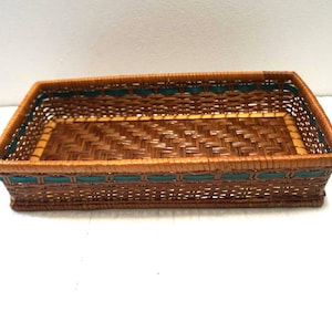 Puede incluir: Cesta rectangular tejida con ribete marrón y verde azulado. La cesta está hecha de material tejido y tiene forma rectangular. El ribete verde azulado añade un toque decorativo a los bordes de la cesta.