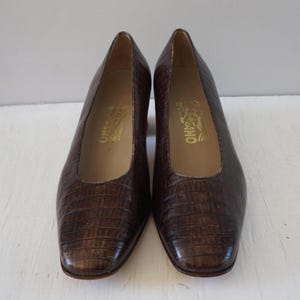 May include: A pair of brown leather pumps with a crocodile embossed pattern. The shoes have a low heel and a square toe. The brand name "Salvatore Ferragamo" is printed on the inside of each shoe.