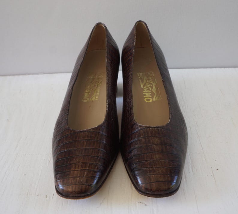 May include: A pair of brown leather pumps with a crocodile embossed pattern. The shoes have a low heel and a square toe. The brand name "Salvatore Ferragamo" is printed on the inside of each shoe.