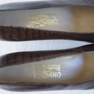 May include: A pair of brown leather shoes with a crocodile embossed pattern. The shoes have a pointed toe and a low heel. The inside of the shoes are lined with a light brown fabric. The shoes have a gold label with the text "Salvatore Ferragamo" on the inside.