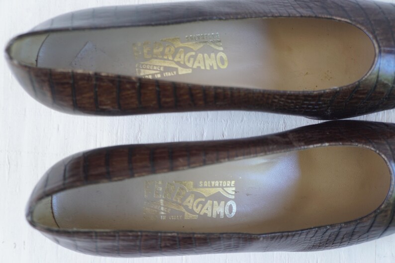 May include: A pair of brown leather shoes with a crocodile embossed pattern. The shoes have a pointed toe and a low heel. The inside of the shoes are lined with a light brown fabric. The shoes have a gold label with the text "Salvatore Ferragamo" on the inside.