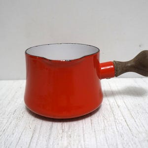Puede incluir: Cacerola pequeña de esmalte rojo con asa de madera. El interior es blanco y el asa es de color marrón oscuro. La cacerola está diseñada para su uso en la estufa y probablemente sea vintage.