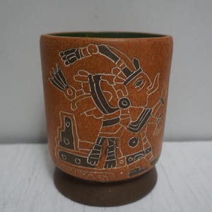 Könnte beinhalten: Ein brauner Keramikbecher mit dunkelbraunem Boden und grünem Innenraum. Die Außenseite zeigt eine weiß-braune Illustration einer Figur im antiken Stil. Der Becher wird wahrscheinlich zum Trinken verwendet.