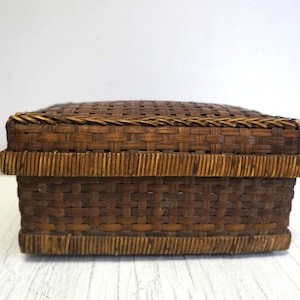 May include: A rectangular, woven basket with a lid. The basket is made of dark brown and tan-colored woven material. The lid and base have a decorative border. The basket is likely used for storage or decoration.