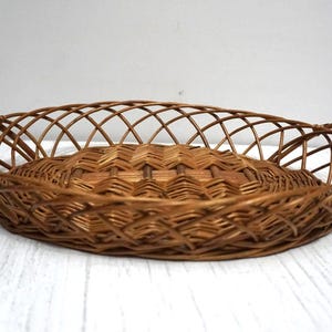 May include: Oval-shaped brown wicker basket with woven base and lattice-style sides. The basket has two curved handles and is suitable for serving or storage. The basket is made of natural materials.