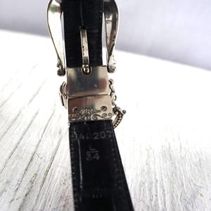 K&ouml;nnte beinhalten: Nahaufnahme eines schwarzen Lederg&uuml;rtels mit einer silbernen Schnalle. Die Schnalle hat ein verziertes Design. Der G&uuml;rtel ist mit L 34 gekennzeichnet, was einer Gr&ouml;&szlig;e von 86 cm entspricht.