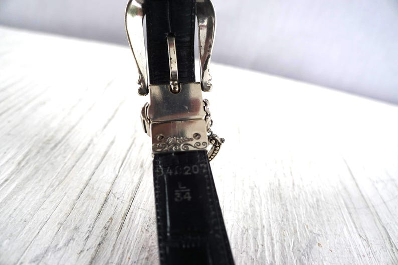 K&ouml;nnte beinhalten: Nahaufnahme eines schwarzen Lederg&uuml;rtels mit einer silbernen Schnalle. Die Schnalle hat ein verziertes Design. Der G&uuml;rtel ist mit L 34 gekennzeichnet, was einer Gr&ouml;&szlig;e von 86 cm entspricht.