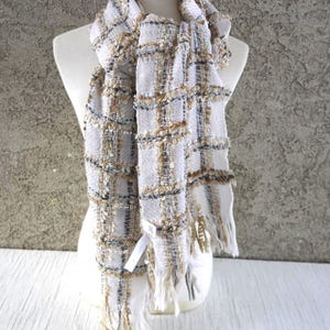 May include: A woven scarf with a grid pattern in white, tan, and black. The scarf has a fringed edge and is draped over a mannequin. The fabric has a textured appearance with subtle sparkle.