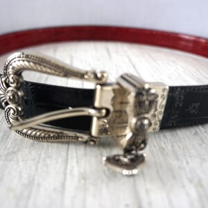 K&ouml;nnte beinhalten: Schwarzer G&uuml;rtel mit roter Innenseite und silberfarbener Schnalle. Die Schnalle hat ein verziertes Design mit einem kleinen Anh&auml;nger. Der G&uuml;rtel ist 3,18 cm breit.