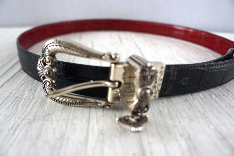 K&ouml;nnte beinhalten: Schwarzer G&uuml;rtel mit roter Innenseite und silberfarbener Schnalle. Die Schnalle hat ein verziertes Design mit einem kleinen Anh&auml;nger. Der G&uuml;rtel ist 3,18 cm breit.