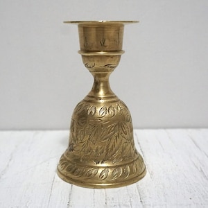 May include: A brass candlestick holder with a bell-shaped base and a detailed floral pattern. The candlestick has a small, circular top to hold a candle. The overall design is ornate and vintage, with a warm, golden hue.