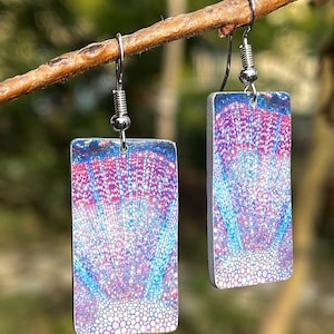 Puede incluir: Un par de pendientes rectangulares con un diseño abstracto y colorido. Los pendientes presentan un patrón de tonos rojos, azules y blancos, que se asemeja a una sección transversal de tejido vegetal. Cuelgan de ganchos plateados.