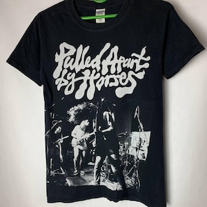 Könnte beinhalten: Schwarzes T-Shirt mit einem weißen Grafikdruck der Band "Pulled Apart by Horses". Die Grafik zeigt ein Bandfoto und den Bandnamen in einer stilisierten Schriftart.