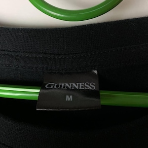 Puede incluir: Camiseta negra con una etiqueta que dice "GUINNESS" y la talla "M". La camiseta est&aacute; hecha de un material suave. Un objeto circular verde est&aacute; en el fondo.