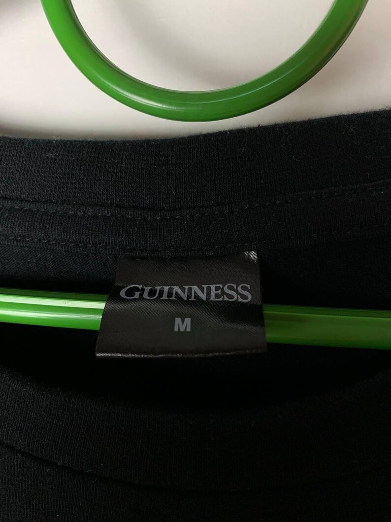 Puede incluir: Camiseta negra con una etiqueta que dice "GUINNESS" y la talla "M". La camiseta est&aacute; hecha de un material suave. Un objeto circular verde est&aacute; en el fondo.