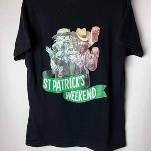 Puede incluir: Camiseta negra con un gr&aacute;fico colorido de cuatro personas sosteniendo jarras de cerveza. El gr&aacute;fico incluye el texto "ST PATRICK'S WEEKEND" en una pancarta verde. La camiseta est&aacute; colgada en una percha verde.