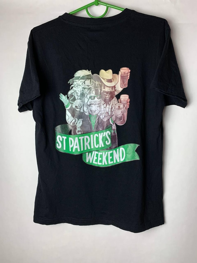 Puede incluir: Camiseta negra con un gr&aacute;fico colorido de cuatro personas sosteniendo jarras de cerveza. El gr&aacute;fico incluye el texto "ST PATRICK'S WEEKEND" en una pancarta verde. La camiseta est&aacute; colgada en una percha verde.