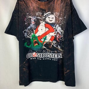 May include: Black t-shirt with a bleached effect featuring a graphic print of the Ghostbusters logo and characters, including Slimer, the Stay Puft Marshmallow Man, and the Ghostbusters logo. The text "Ghostbusters" and "Who You Gonna Call?" is printed on the shirt.