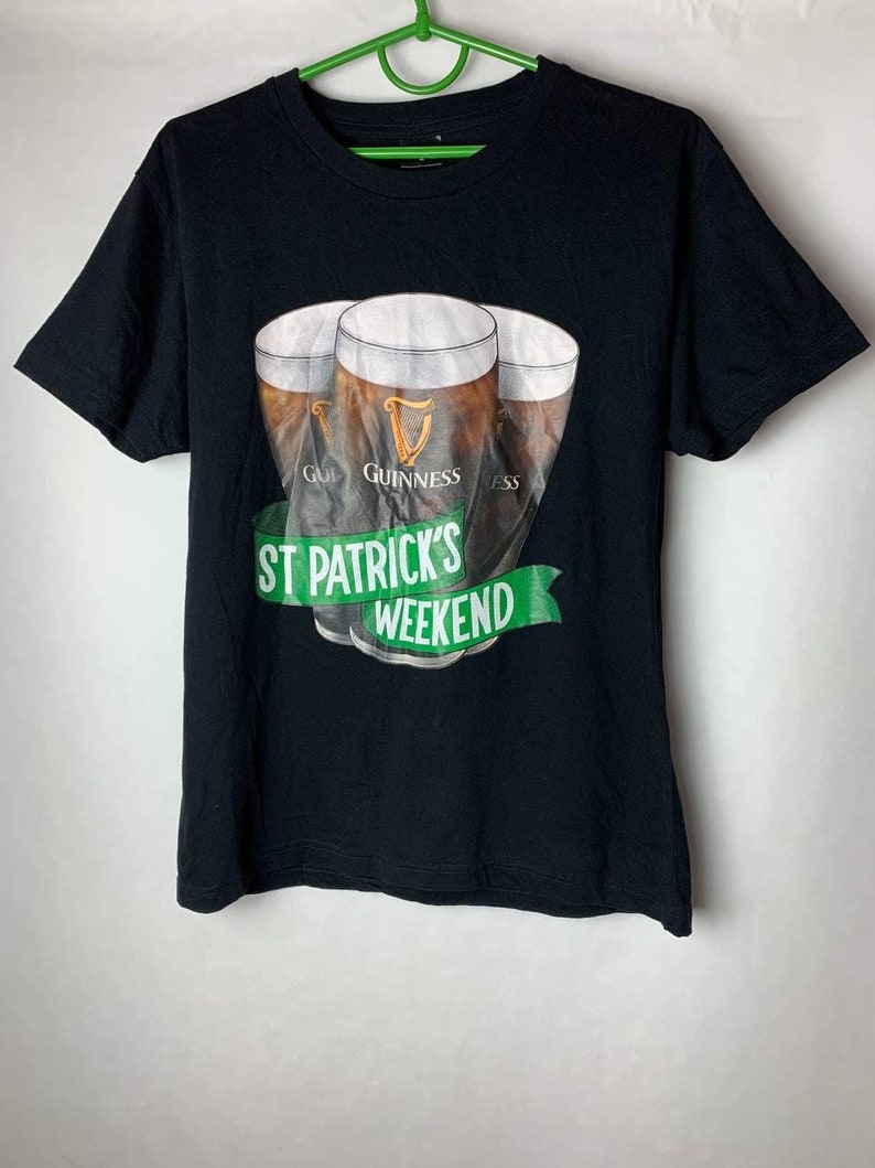 Puede incluir: Camiseta negra con un gr&aacute;fico de tres pintas de Guinness. El logotipo de Guinness es visible. Una pancarta verde debajo dice "St Patrick's Weekend". La camiseta tiene mangas cortas y cuello redondo.
