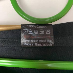 Puede incluir: Primer plano de una etiqueta de ropa negra con texto blanco. La etiqueta dice "100% Algod&oacute;n" e incluye s&iacute;mbolos de cuidado para lavar y planchar. Tambi&eacute;n dice "No planchar sobre la zona estampada" y "Hecho en Bangladesh."
